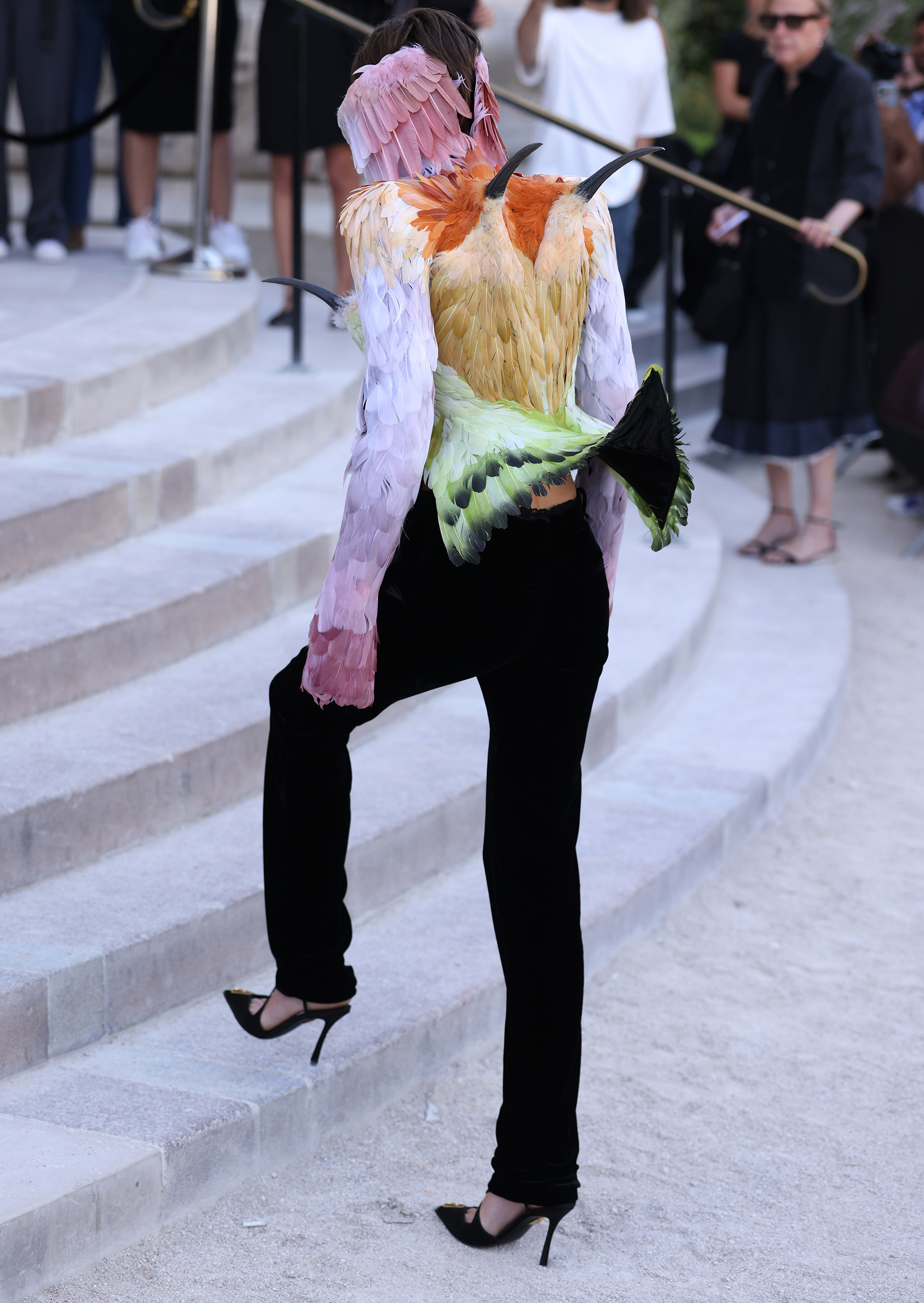 Emma Corrin wears bird blazer with tail in back, as seen from behind while they are walking up stairs at Schiaparelli 2026-2027 show.