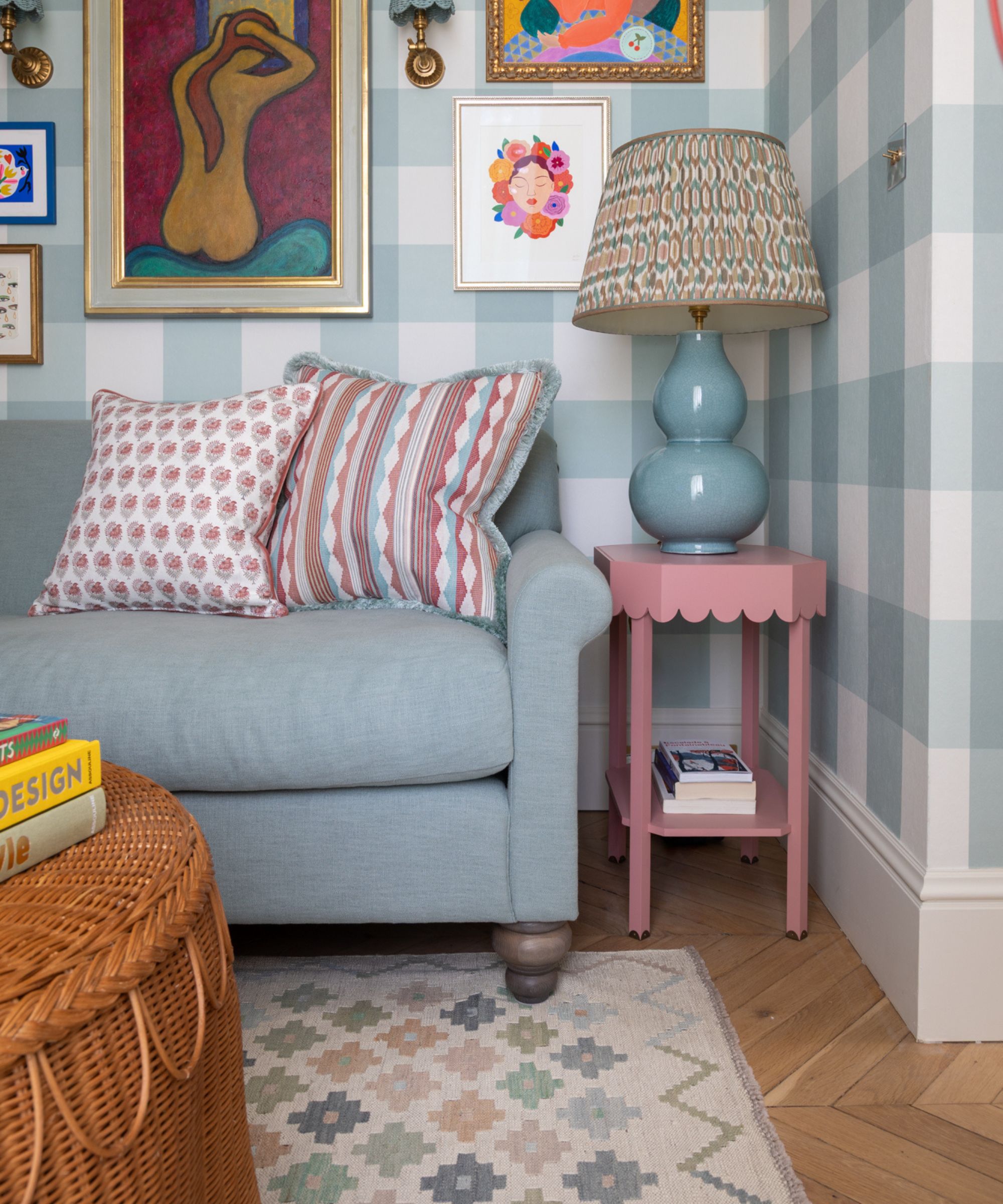 blue gingham/plaid wallpapered snug room with a blue couch, multicolored blue and pink pillows, a pink scalloped side table with a blue lamp, rattan scalloped coffee table sat on a printed aztec rug in pastel shades