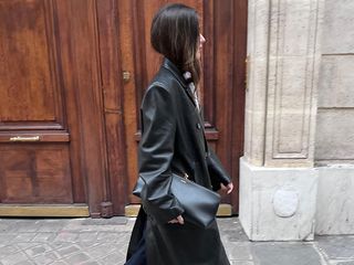 woman wears black trench coat while holding a giant large black clutch and black pumps with tights