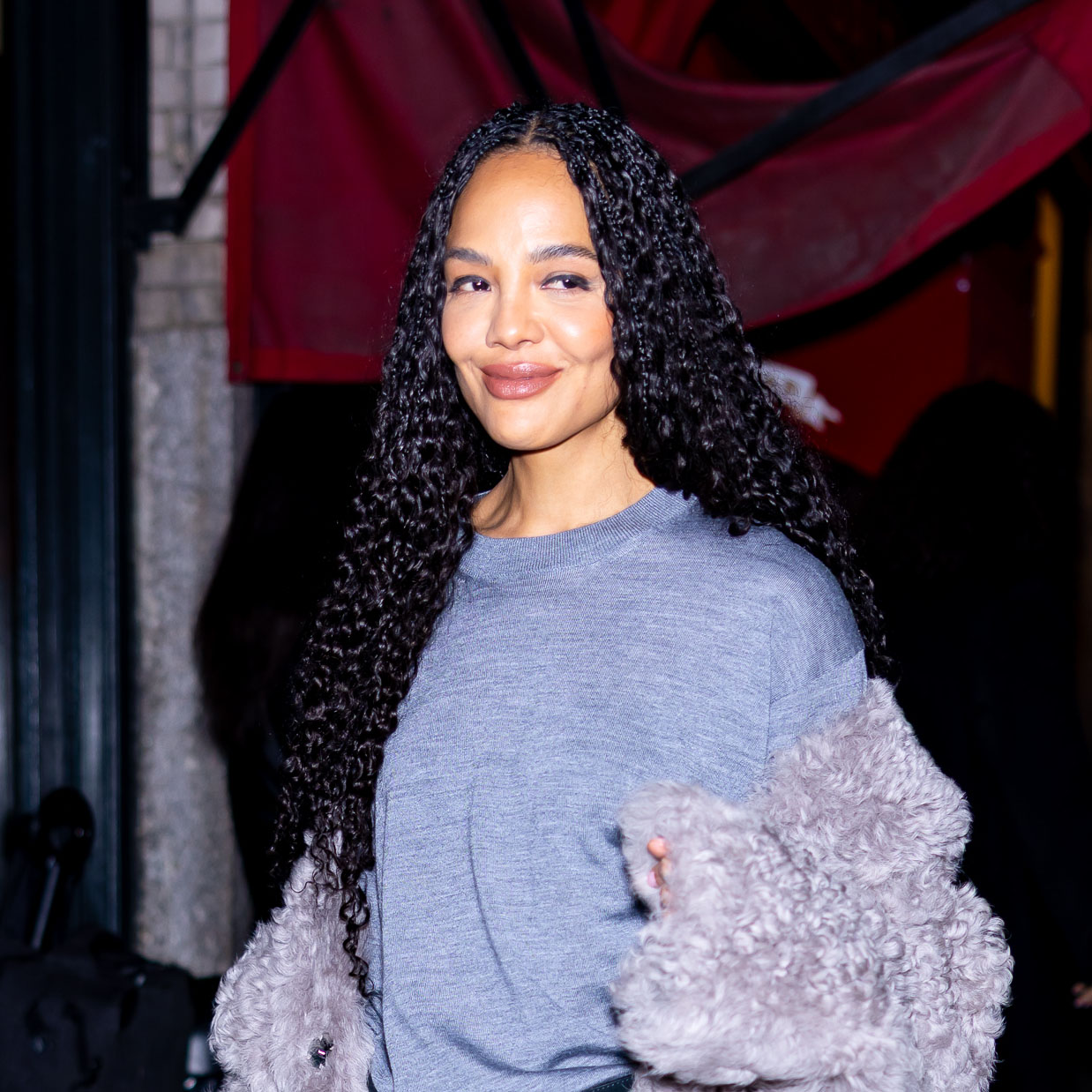 Tessa Thompson wearing a gray sweater, gray fur jacket, black satin skirt, and black boots