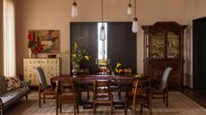 dining room with pale pink walls, timber dining table with timber chairs, rug, sofa on one side of the room, yellow console with vase of flowers and artwork styled above it, and on the other side fo the room is a large timber dresser with glass-front
