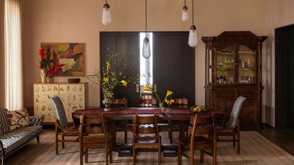 dining room with pale pink walls, timber dining table with timber chairs, rug, sofa on one side of the room, yellow console with vase of flowers and artwork styled above it, and on the other side fo the room is a large timber dresser with glass-front
