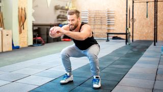 A man performs squats in an empty gym. His knees and hips are bent, hands held out straight before him, and chest upright. Behind him we see barbells and gym rings.