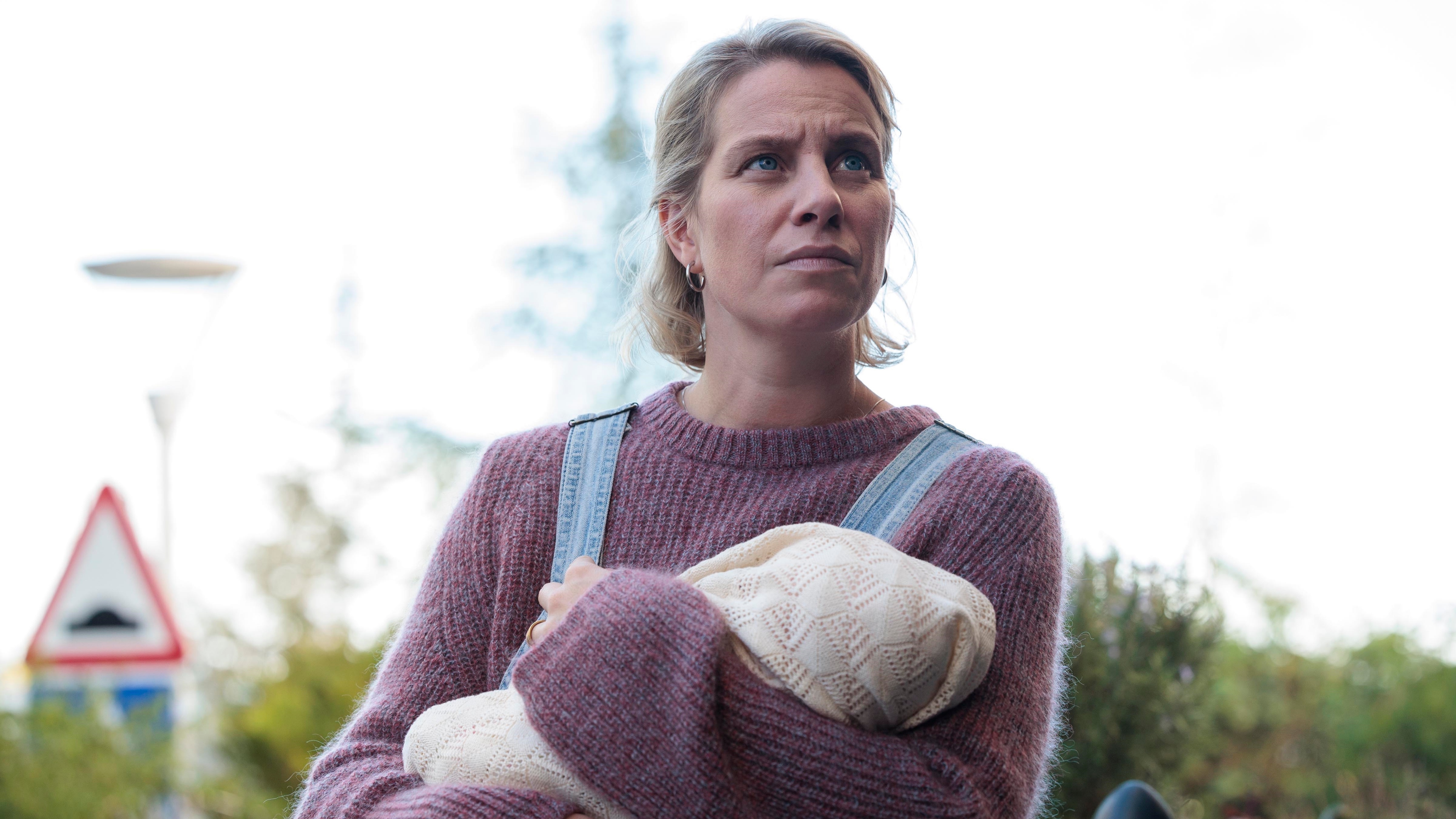 Tina holding a baby and looking at someone off camera with a worried look