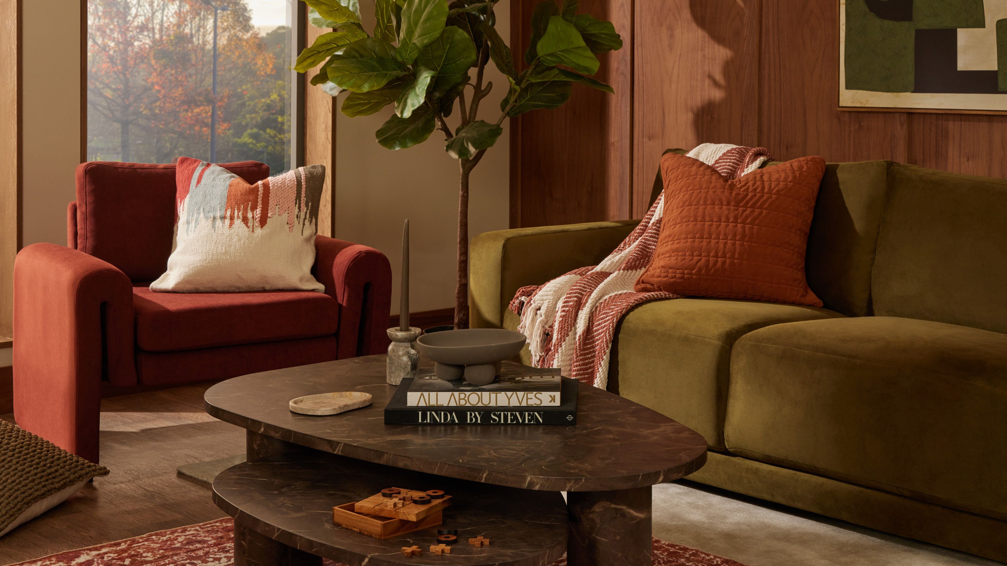 retro-inspired living room with green velvet sofa, with rust red armchair, rust cushion and throw, marble nesting coffee table, red patterned rug, fiddle leaf plant and timber-clad wall with artwork