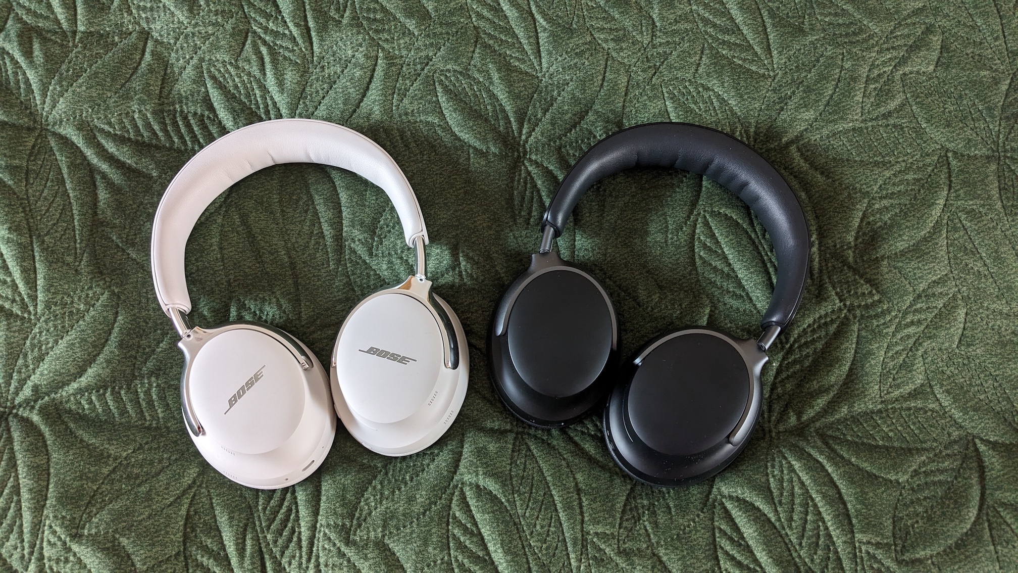 A white pair of Bose QC Ultra Headphones (2nd Gen) lying on a green surface next to their black 1st Gen predecessors.