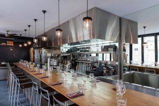 The kitchen bar of an industrially decorated restaurant features warm lights hanging from the ceiling, a wooden counter and tall chairs.