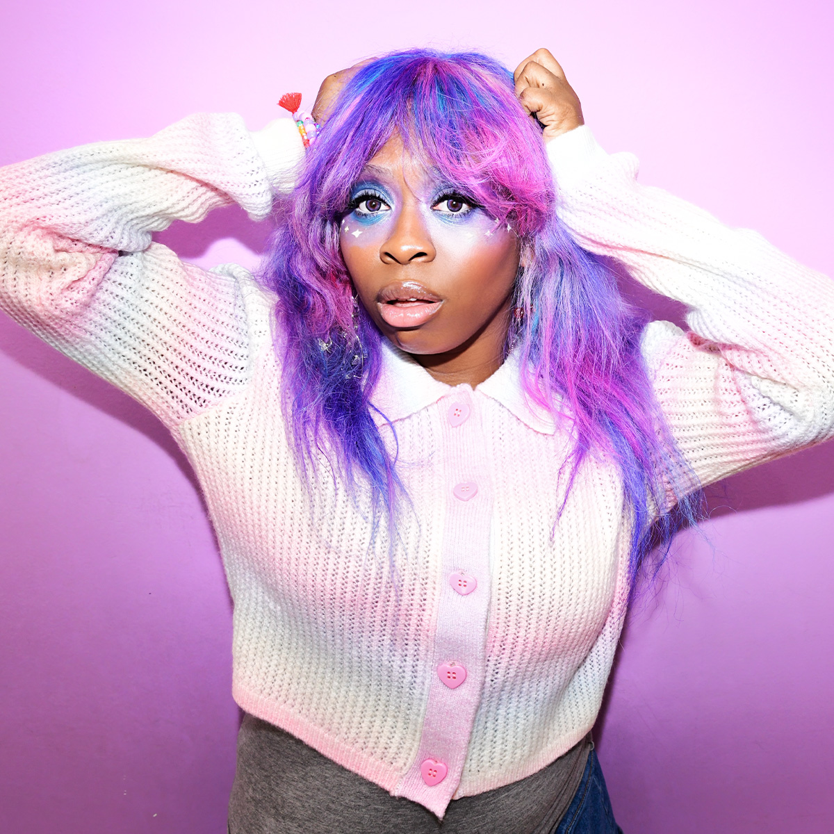 Artist hemlocke springs is photographed against a purple backdrop. She is wearing a rainbow-colored cardigan with pink, heart-shaped buttons, and her hair is bright pink and purple.