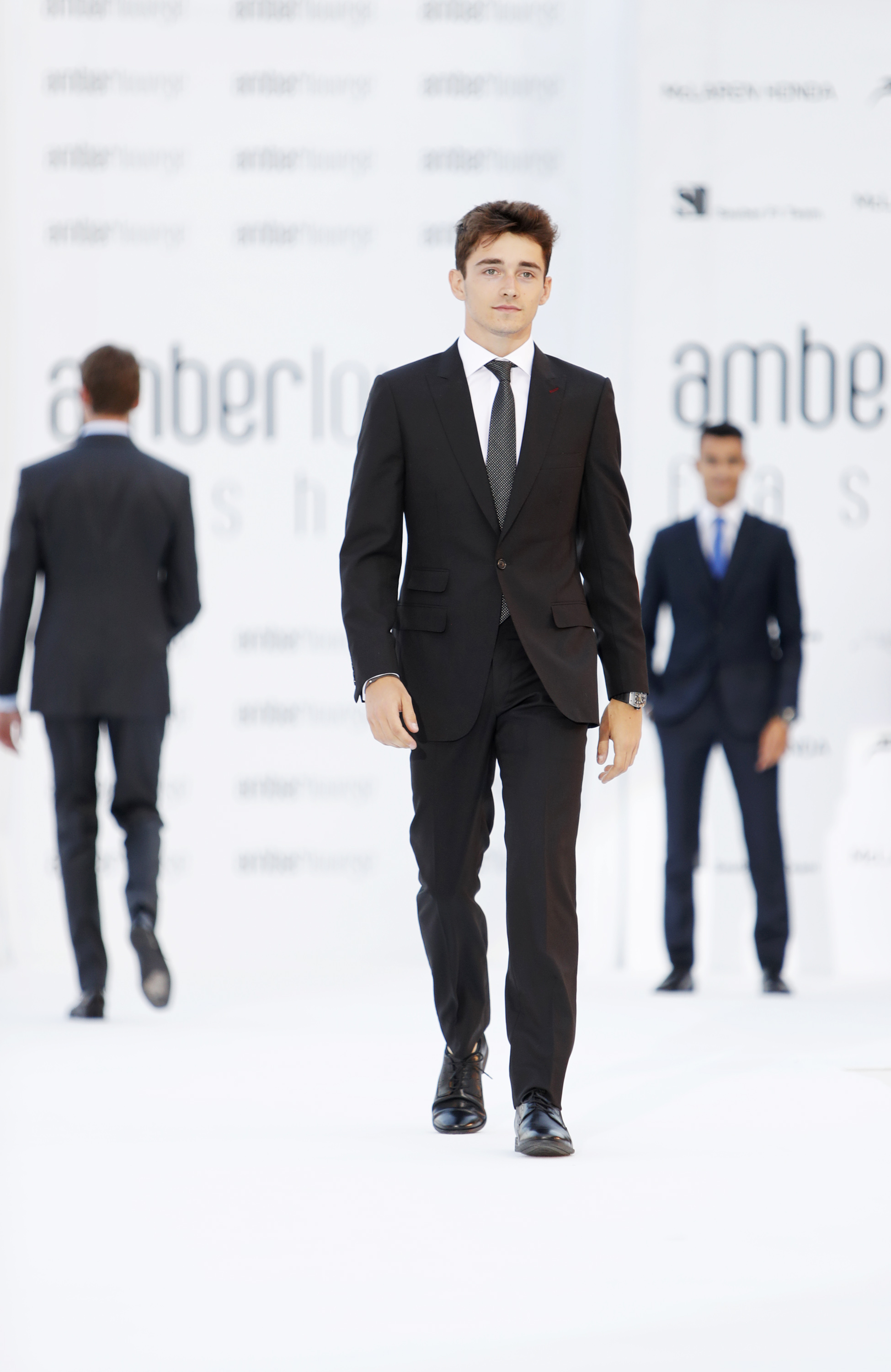 Charles Leclerc walks down a white catwalk in a black suit and tie