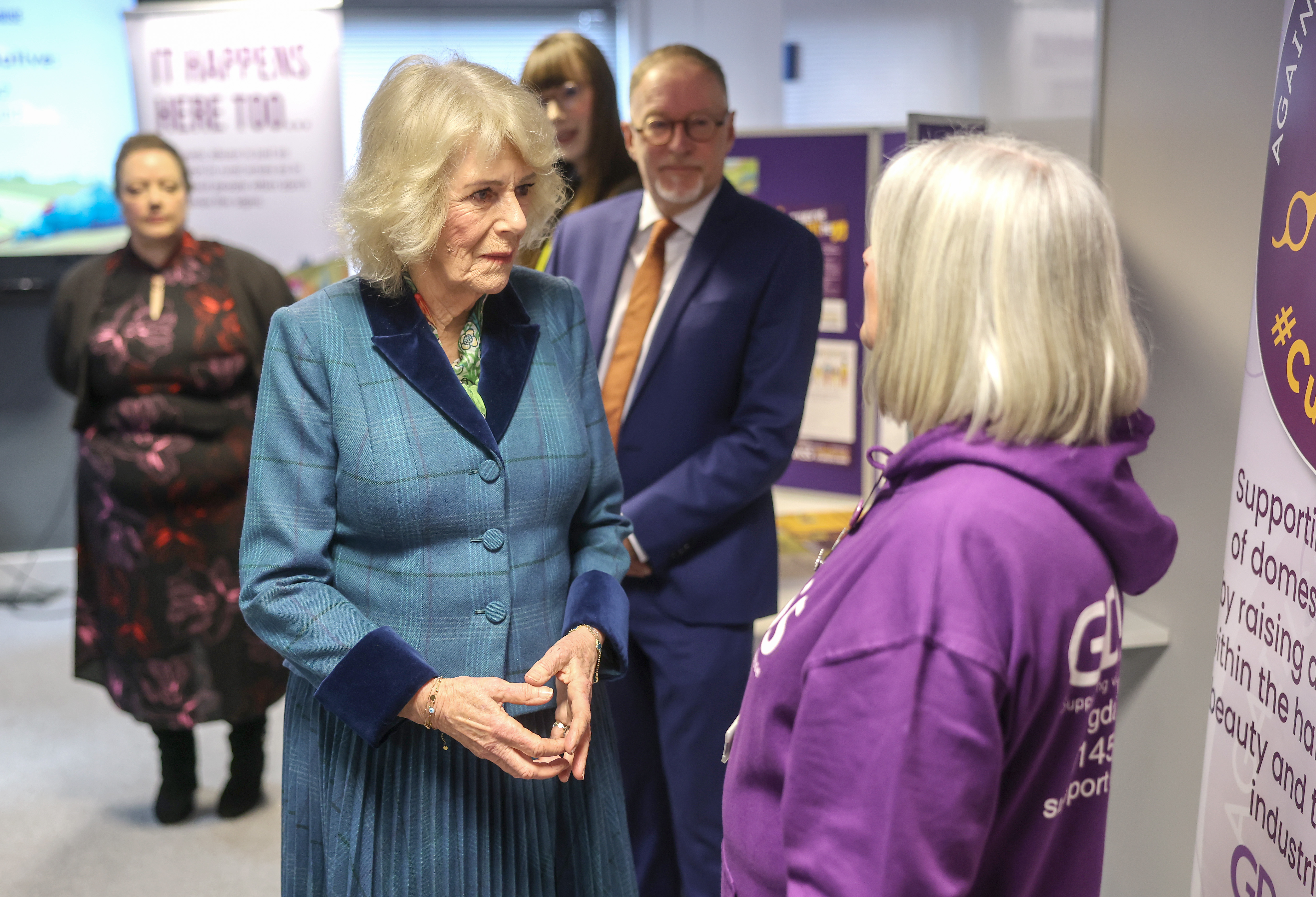 Queen Camilla wearing a blue suit talking to a woman in a purple hoodie