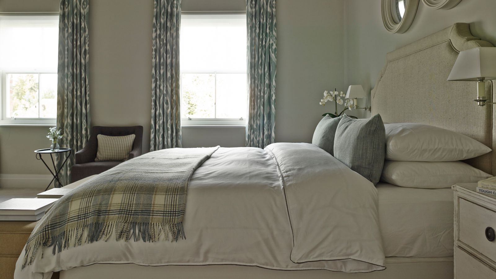 A neutral bedroom with a large bed to the right and in the foreground, with white bedding, blue cushions, and a plaid blanket. The walls are cream, and on the back wall there are two large windows with blue patterned curtains. Above the bed are two circular white mirrors.