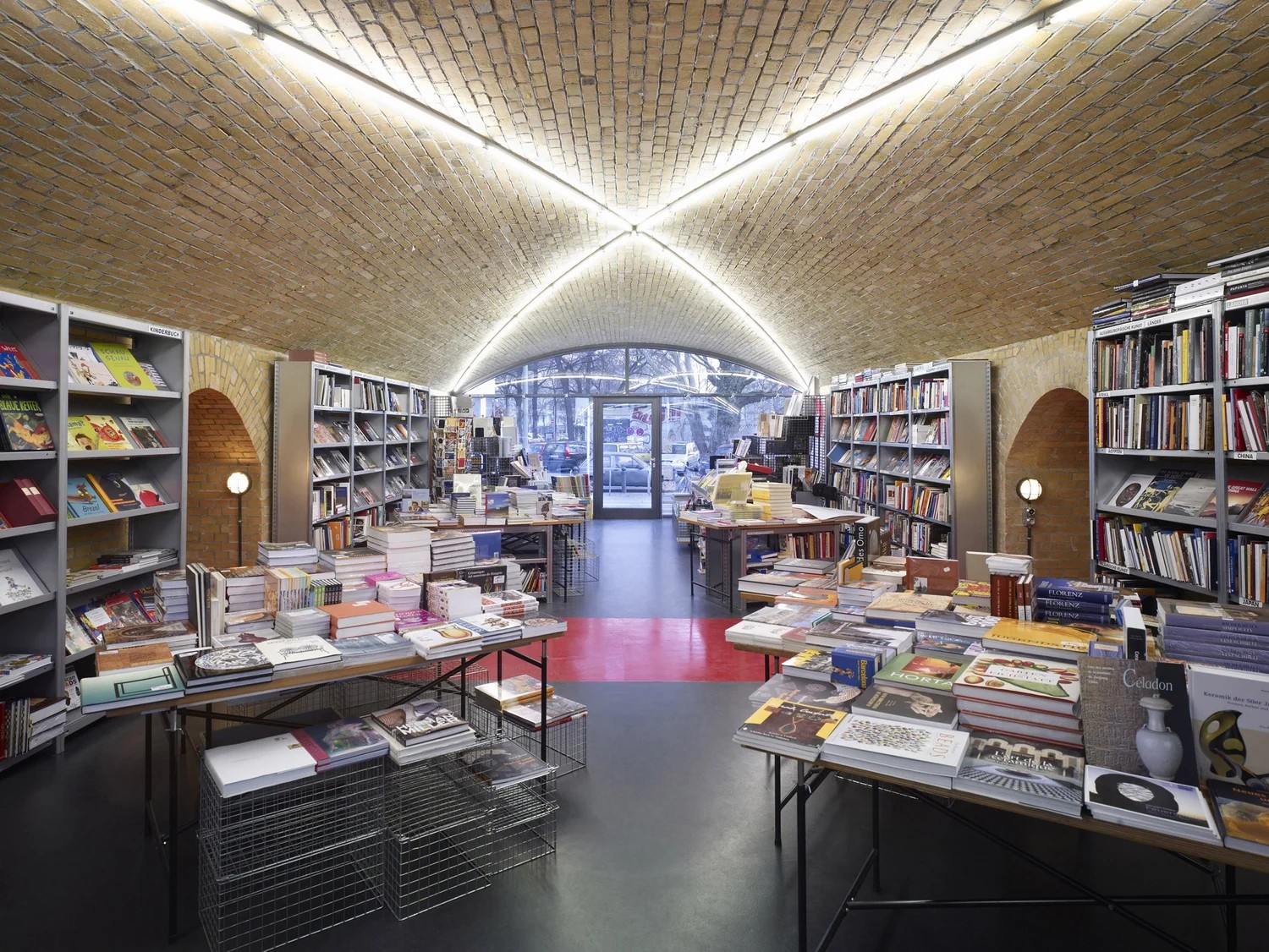 A beautifully designed bookshop with vaulted brick walls lit by neons, chrome furniture and shelving, and a cement black and red floor.