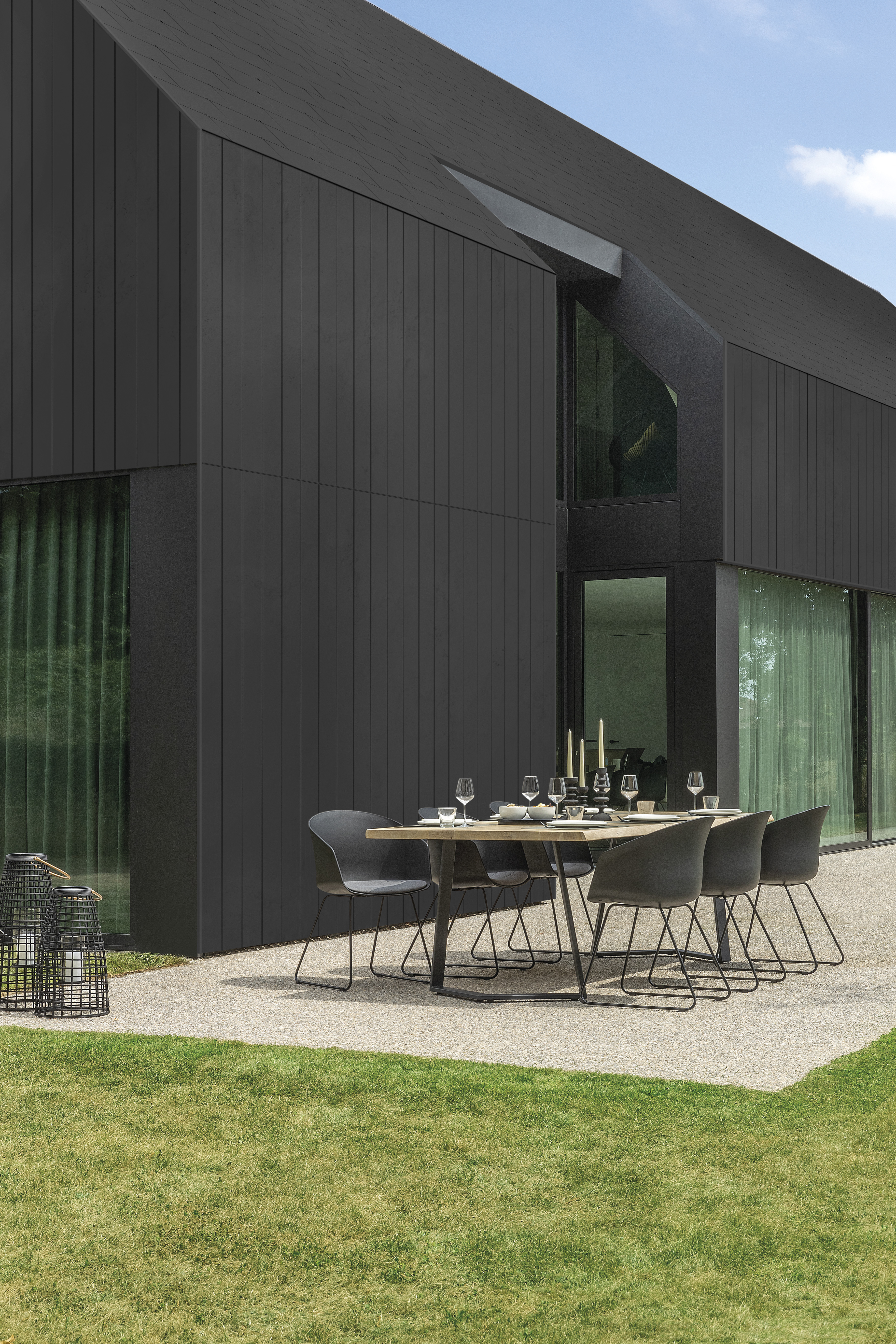 Image of the exterior of a house covered in black fibre cement cladding. There is an outdoor dining table set with glassware and decor and six black dining chairs around it.