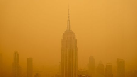 photo shows the empire state building as viewed from a nearby skyscraper; the building and those nearby are surrounded by a dense cloud of yellow-orange smog made up of wildfire smoke