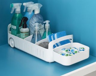 YouCopia white under sink organizer caddy with wheels containing an assortment of cleaning products and dishwasher tablets