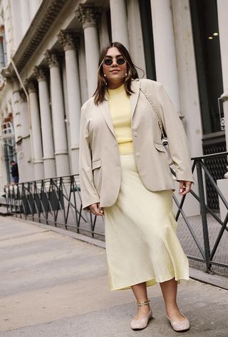 A woman wearing a tan blazer over a yellow sweater with a yellow slip skirt and mesh ballet flats.