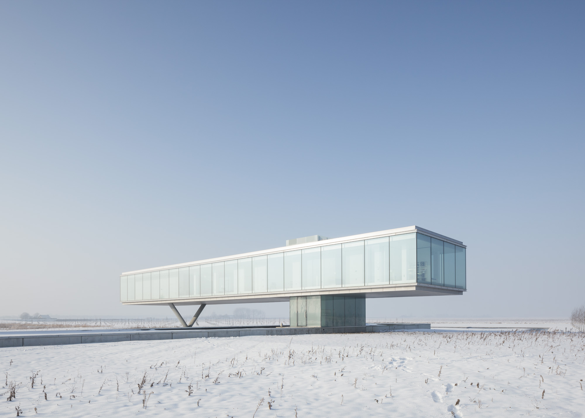 A flat and wide building of concrete, metal and glass dutch house stands alone in a field of snow. 