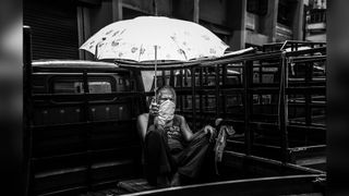 Black-and-white image of a man holding an umbrella while sitting in the back of a van, wearing a COVID mask.