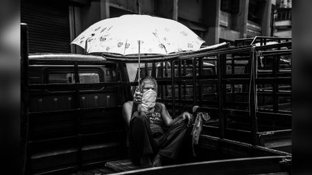 Black-and-white image of a man holding an umbrella while sitting in the back of a van, wearing a COVID mask.