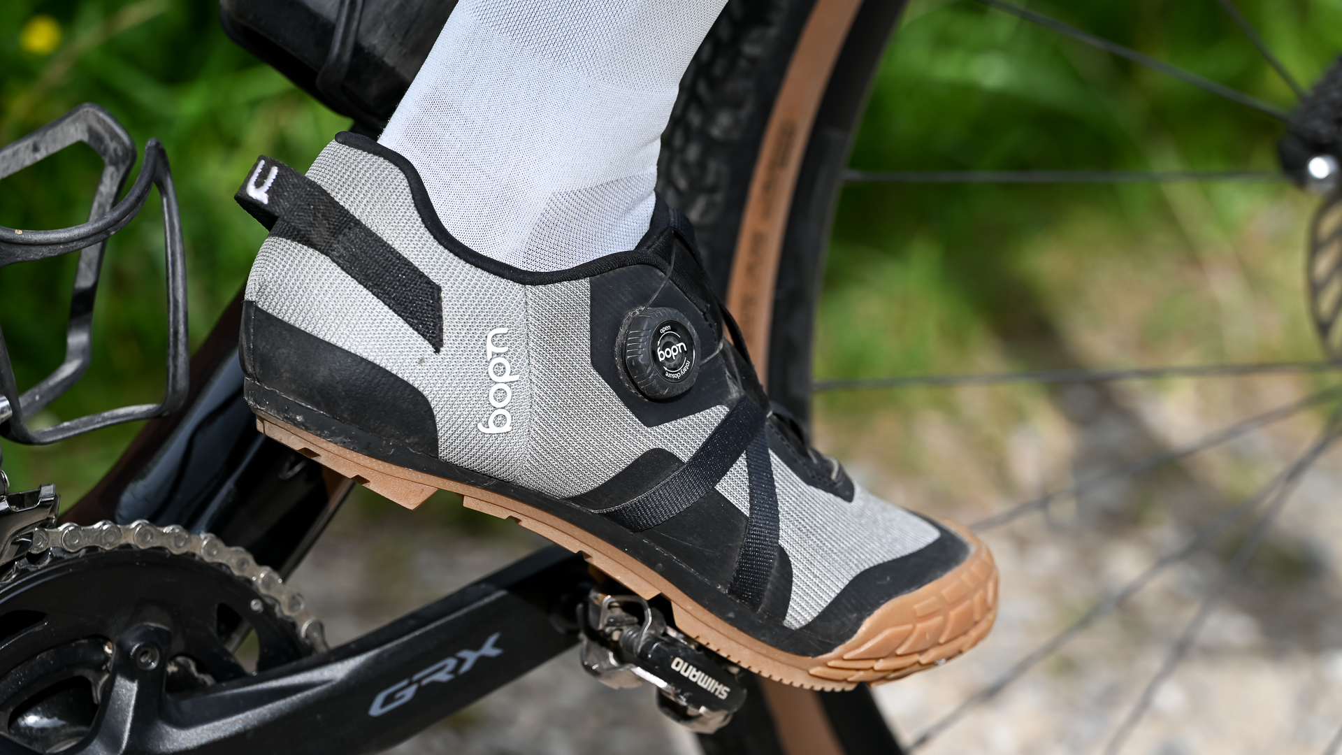 Close up of a foot wearing white socks and grey shoes clipped into a pedal on a black gravel bike