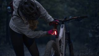 Low-key photo of a cheerful young woman with her enduro mountain bike (eMTB) at night, turning on the rear light.