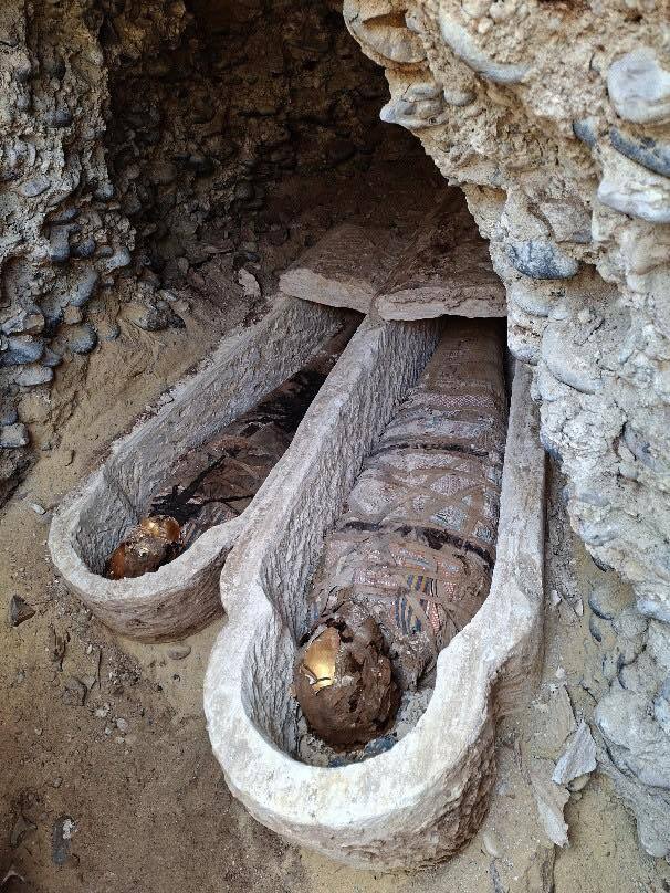 Two mummies lie in their coffins inside a cylindrical tunnel in the wall. Their heads are toward the camera