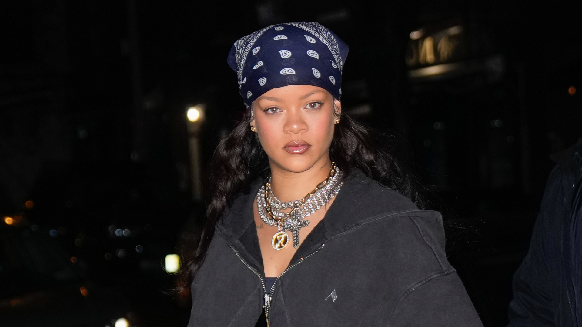 A woman wearing a bandana hair scarf wearing a zip up hoodie, multiple diamond chains, and lipgloss