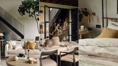 A neutral living room with a cream sofa and orange tone cushions / A dark wood dining table with a vase of stems / A cozy neustral bed with orange cushion, with a small side table