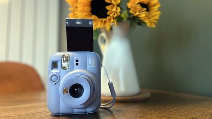 The Frost Blue Instax Mini 13 on a table with a print sticking out of the top of it next to a vase of sunflowers 