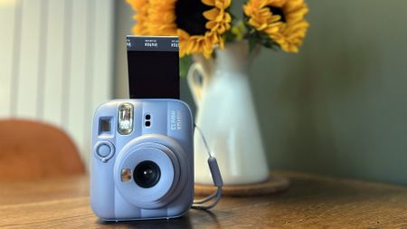 The Frost Blue Instax Mini 13 on a table with a print sticking out of the top of it next to a vase of sunflowers 