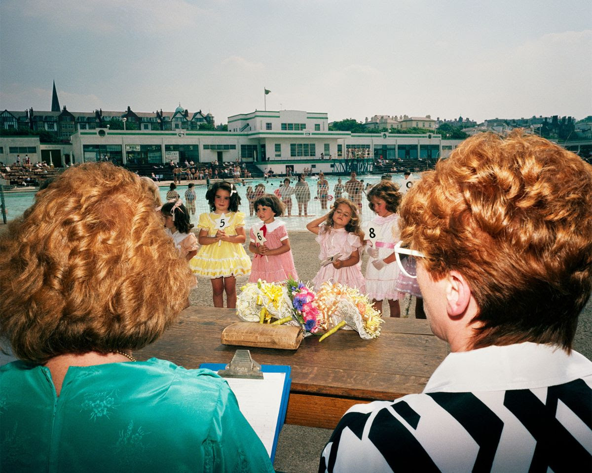 From a behind-the-shoulder perspective of two judges, several young girls in frilly dresses hold numbered signs during a beauty pageant at an outdoor pool.