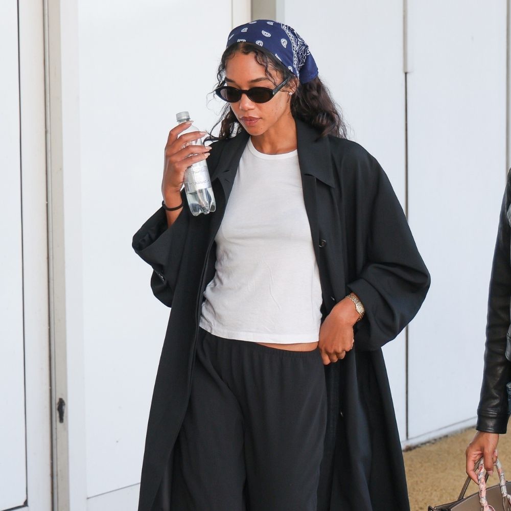 Laura Harrier walks through the airport wearing a white t-shirt, black pull-on trousers and black trousers. She layered with a black trench coat and wears a bandana over her hair.