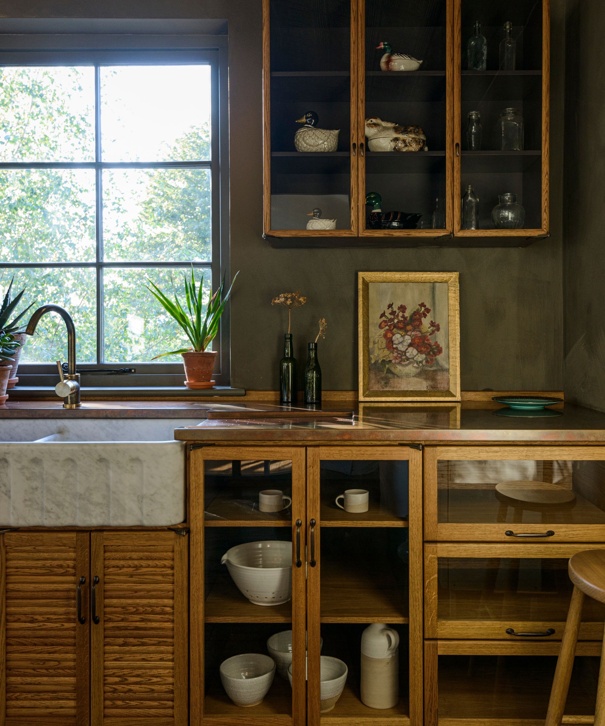 Rustic kitchen corner with wooden cabinets, a white marble farmhouse sink, a copper countertop with a floral painting, and a glass cabinet showing decorative ducks