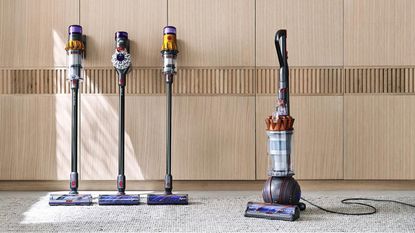 Three Dyson cordless vacuums and one upright Dyson vacuum on a low pile white and gray mottled carpet against wooden cabinets