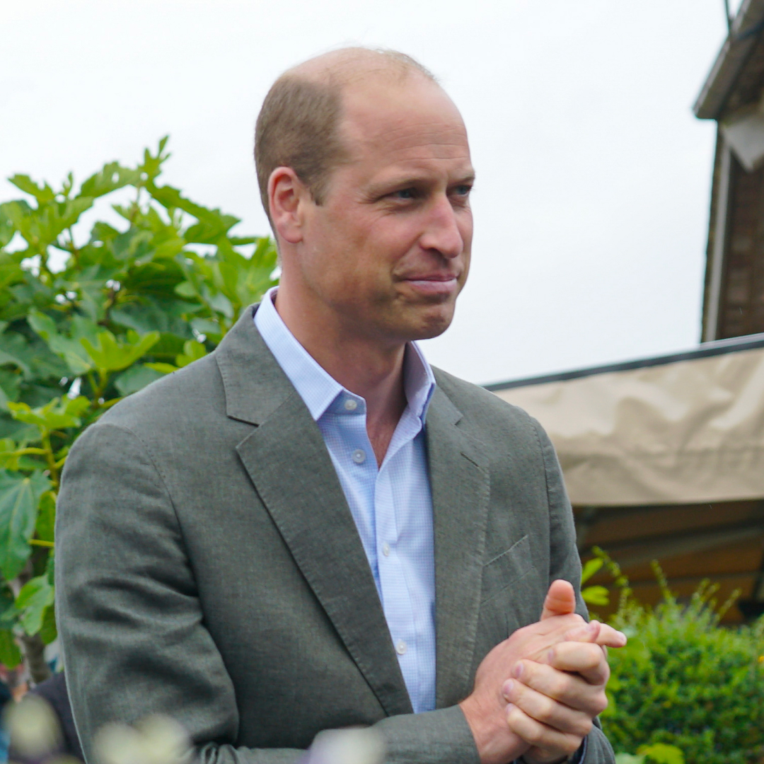 Prince William wears a gray suit and light blue tie and holds his hands together while looking pensive