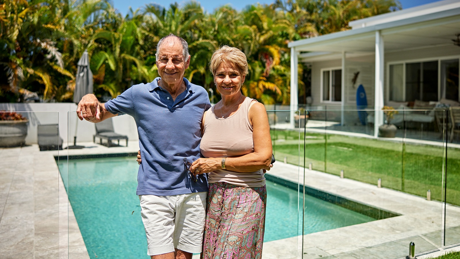Front view of man and woman in 60s and 70s, casually dressed, arms around each other, standing in backyard of home with pool and smiling at camera.