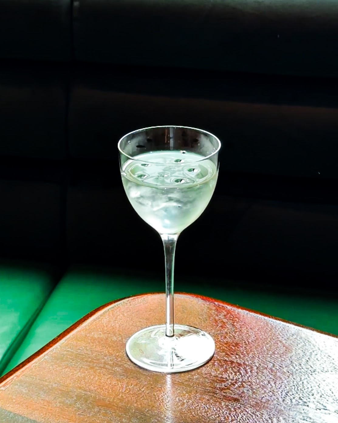 A transparent cocktail glass filled with a transparent drink and ice cubes sits atop a textural, lava stone table with round edges.