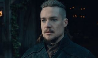 The Last Kingdom Season 5: A battle-scarred Uhtred of Bebbanburg (Alexander Dreymon) stands in dark clothing with his back to a darkened doorway, looking toward the camera with a foreboding expression on his face