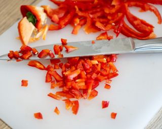 A silver knife resting on a white chopping board with a chopped red bell pepper on a light wooden surface