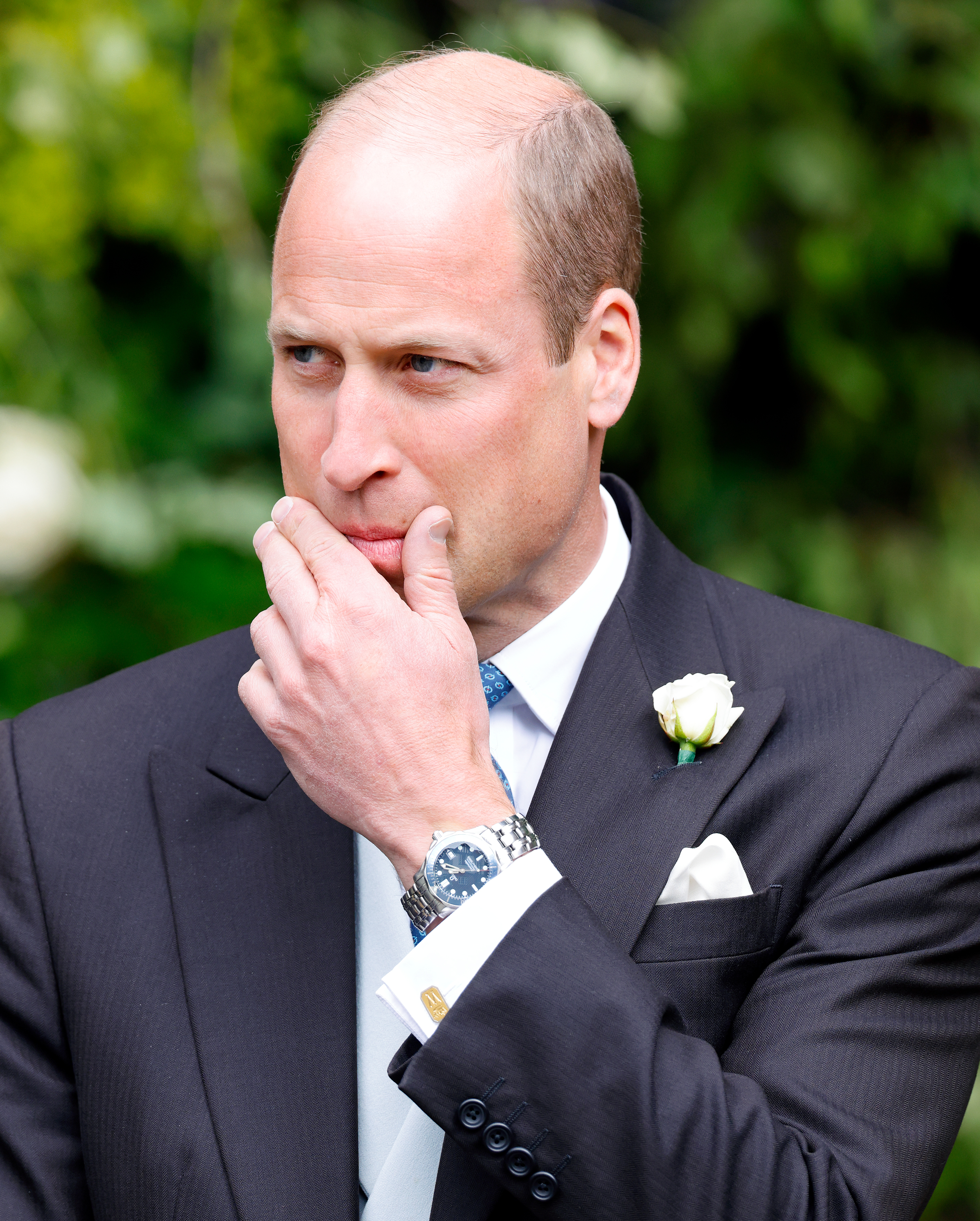 Prince William touching his face, wearing a suit with a white flower pinned to it