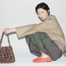 Model crouching holding a brown handbag wearing orange ballet flats a green tartan patchwork skirt and a beige top sold at SSENSE