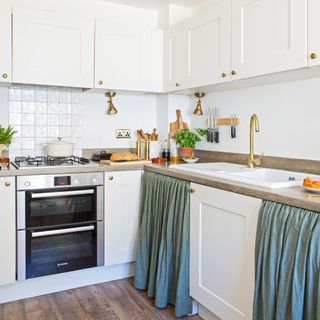 a white kitchen with Shaker units, a built-in oven, brass details, and lighting and blue/green cabinet skirts