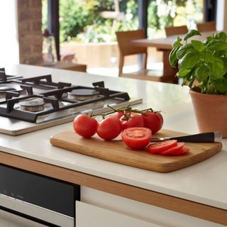 Close up of a wooden chopping board with tomatoes and tomato slices beside a pot of basil and gas hob