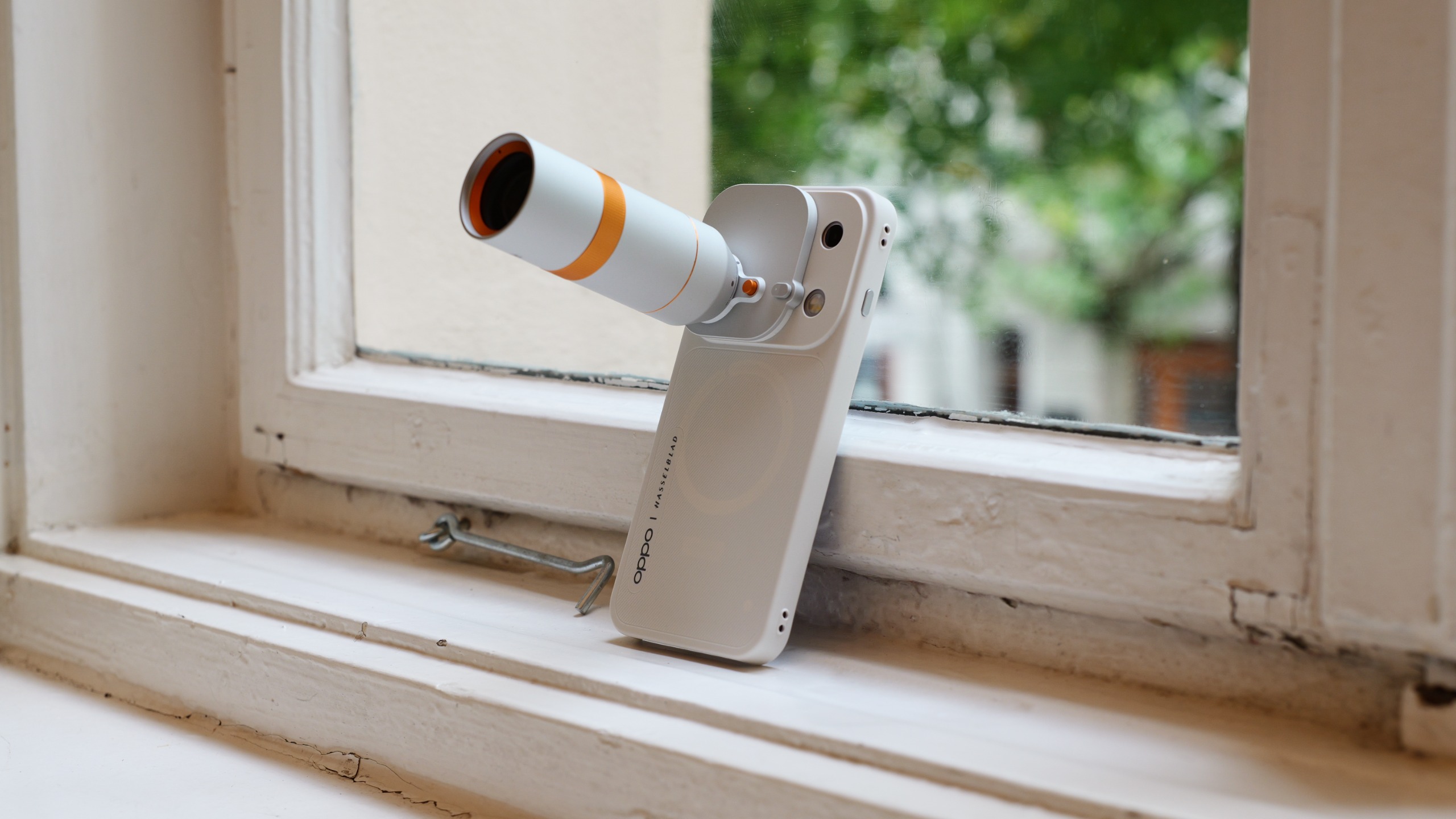 Oppo Find X10 Pro Max standing on a windowsill with its telephoto lens accessory attached, photographed against a softly blurred outdoor background.