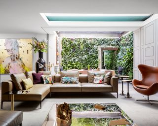 large open plan living room space with leather egg chair and leather large sofa, mirrored square coffee table, large floor to ceiling storage units with slim paneling, outdoor-indoor courtyard space with green living wall, large cream rug and colorful artwork on back wall