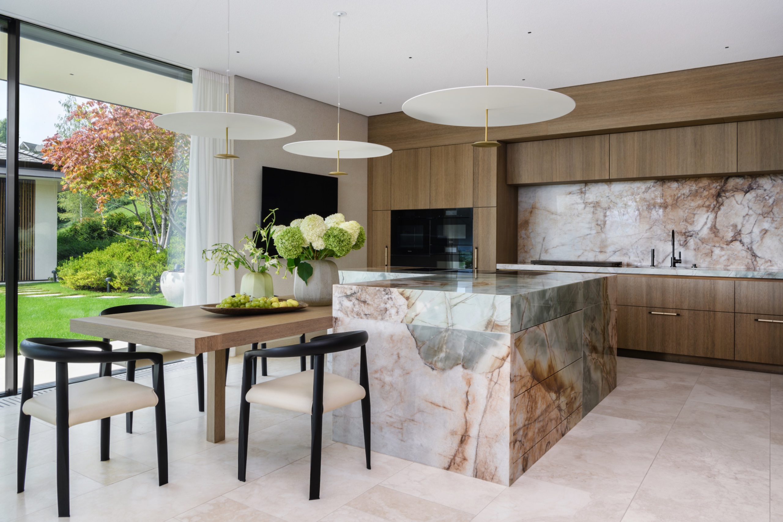 Veined marble kitchen island with an integrated lower wooden dining table and black-framed chairs, surrounded by wood cabinetry and full-height garden windows.
