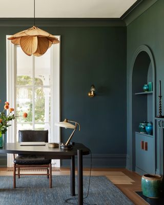 Image of a home office that is color-drenched in a phthalo green paint color. There is a tall, skinny window on the back wall with a black desk and desk chair positioned in front of it. A rattan pendant light hangs above the desk and there is a vase of orange flowers on the desk.