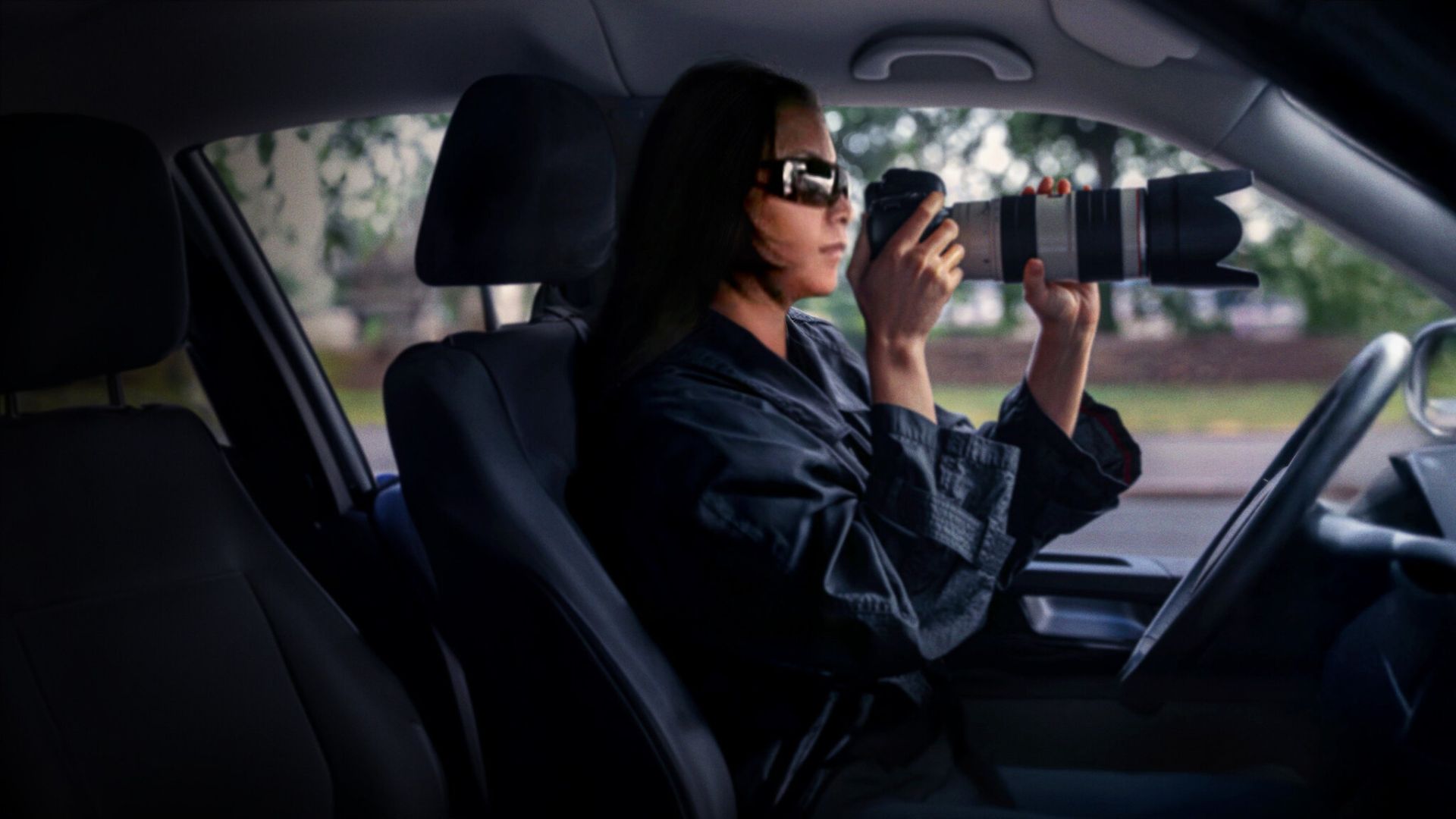 A woman sits in a car wearing sunglasses and holding a camera
