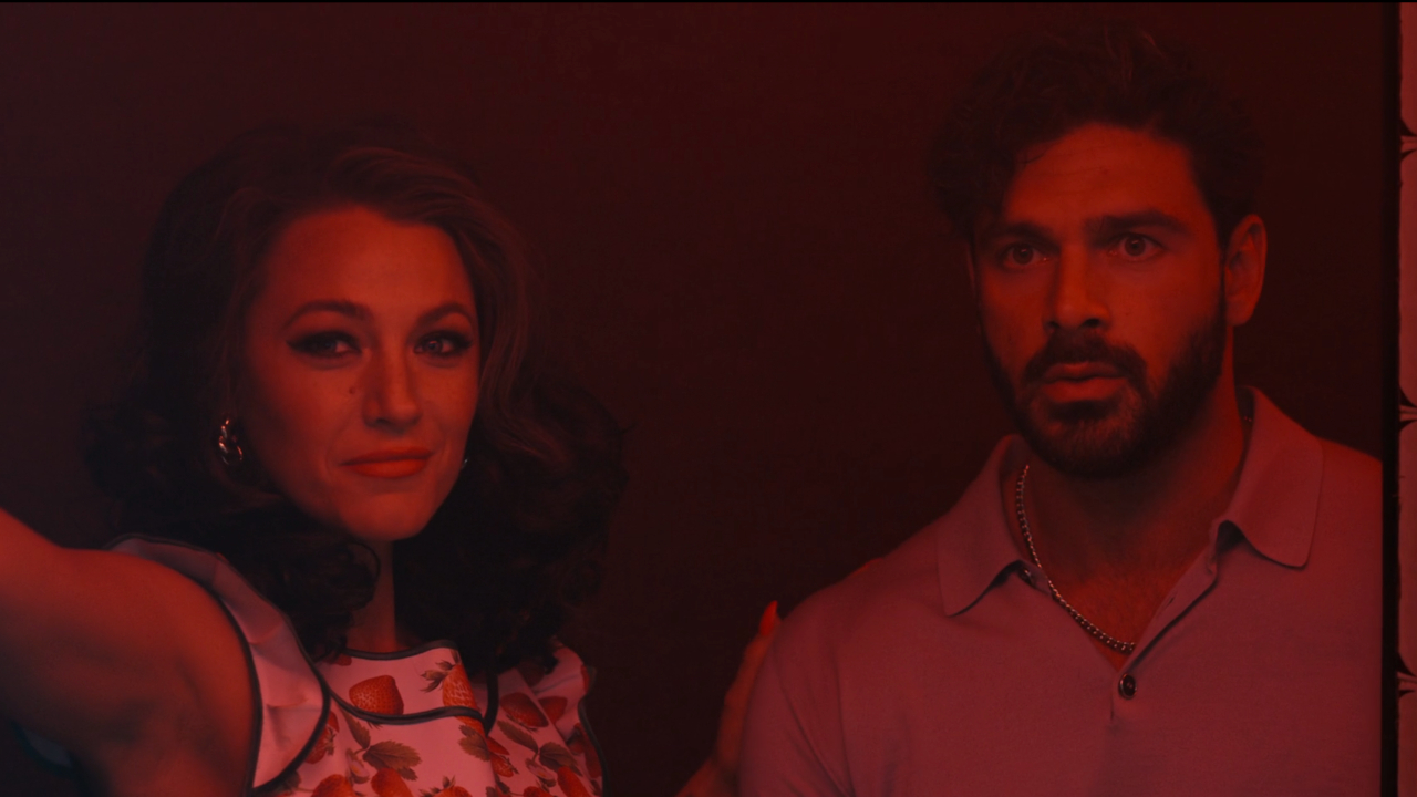 Blake Lively stands smiling next to Michele Morrone in a red lit scene,. in Another Simple Favor.