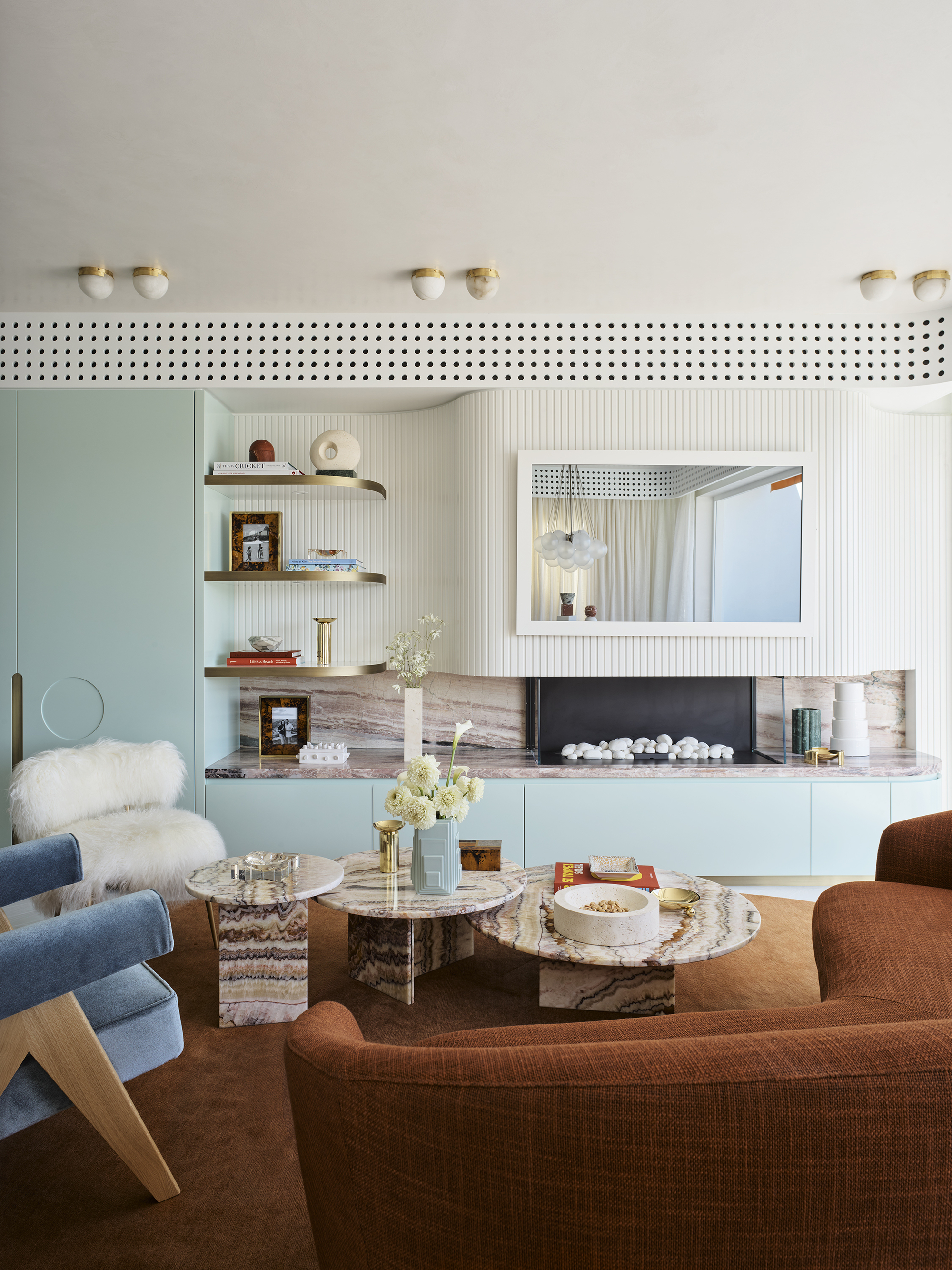 70s style living room with rust orange sofa, orange rug, mint green cabinetry, fluted surface above marble fireplace with mirror/tv, shaggy armchair, blue velvet armchair, perforated detail along the roofline concealing aircon